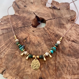 May include: A gold necklace with a hammered metal pendant and turquoise, green, and white beads. The necklace is on a wooden surface.