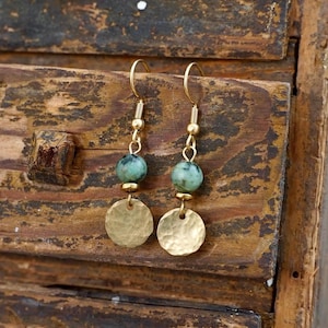 May include: A pair of gold-coloured dangle earrings. Each earring features a gold-coloured hook, a small gold bead, a green stone bead, and a hammered gold disc. The earrings are displayed against a rustic wooden surface.