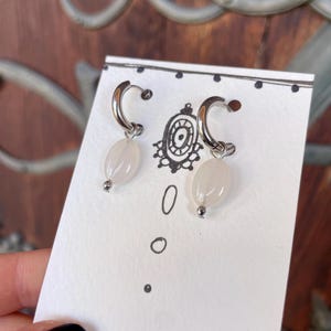 May include: A pair of silver hoop earrings with white oval-shaped beads hanging from them. The earrings are displayed on a white card with black dots.