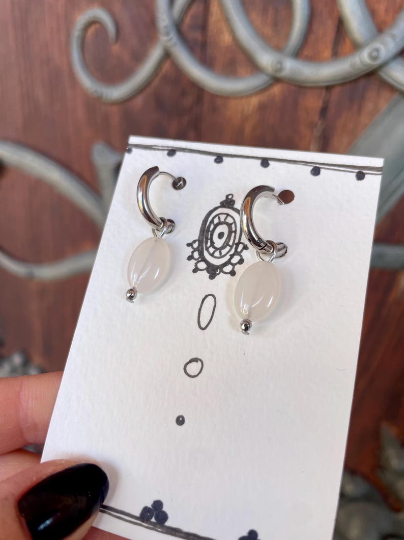 May include: A pair of silver hoop earrings with white oval-shaped beads hanging from them. The earrings are displayed on a white card with black dots.