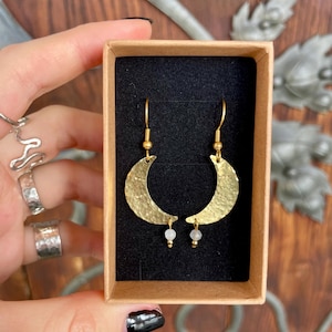 May include: Gold crescent moon earrings with small white beads hanging from the bottom. The earrings are in a small brown box.