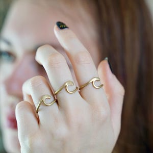 May include: Three gold wave rings worn on a person's fingers. The rings are simple and delicate, with a curved wave design.