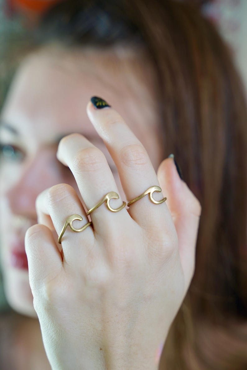 May include: Three gold wave rings worn on a person's fingers. The rings are simple and delicate, with a curved wave design.