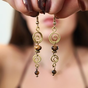 Può includere: Un paio di orecchini pendenti con spirali in filo color oro e perline sfaccettate in occhio di tigre marrone. Ogni orecchino è composto da una spirale superiore, una perla più grande, una seconda spirale e una piccola perla sul fondo.