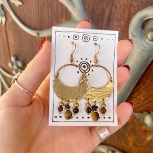 Può includere: Orecchini a cerchio color oro con un design a mezzaluna martellata e perline marroni e dorate pendenti. Gli orecchini sono esposti su una carta bianca con segni decorativi neri.