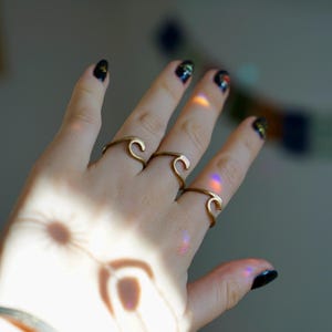 May include: A hand wearing three gold wave-shaped rings. The rings are stacked on two fingers. The hand is against a light background.