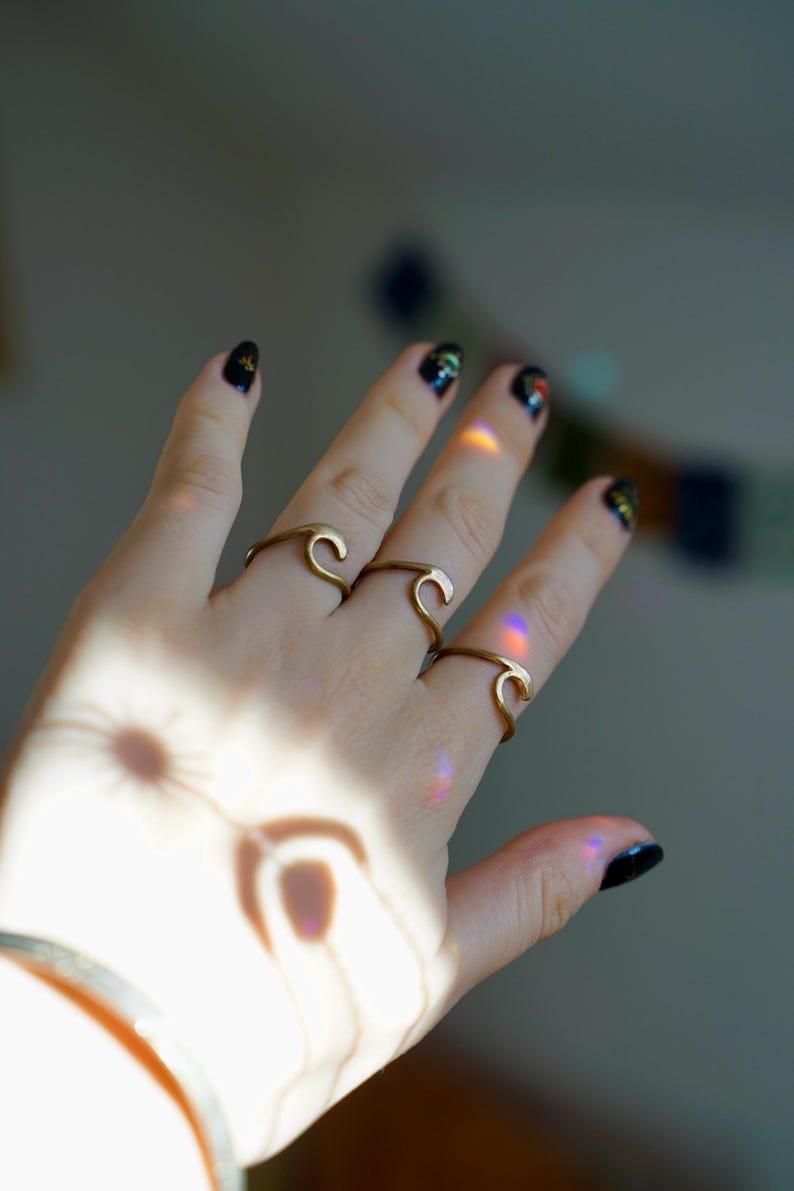May include: A hand wearing three gold wave-shaped rings. The rings are stacked on two fingers. The hand is against a light background.