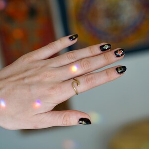 May include: A hand with black nail polish and gold accents on the nails, wearing a gold ring shaped like a wave.