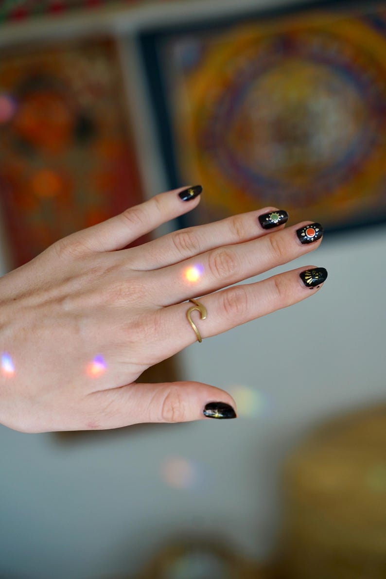 May include: A hand with black nail polish and gold accents on the nails, wearing a gold ring shaped like a wave.