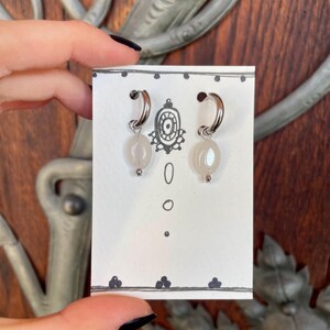 May include: A pair of silver hoop earrings with white oval-shaped beads hanging from them. The earrings are displayed on a white card with black dots.