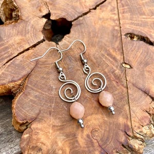 May include: Silver-tone spiral earrings with peach-coloured beads. The earrings are displayed on a textured, brown wooden surface. The earrings have a hook closure.
