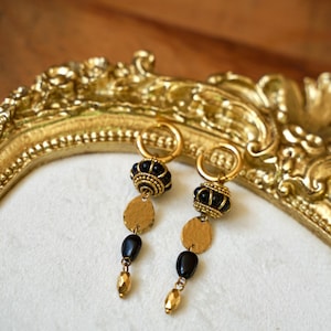 May include: Gold hoop earrings with black beads and gold discs. The earrings are hanging from a gold frame.