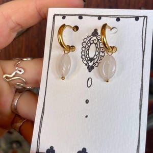 May include: A pair of gold hoop earrings with a white stone pendant hanging from each hoop.