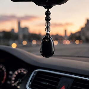 Op de afbeelding: Een hangend ornament voor de achteruitkijkspiegel van een auto met een gladde, gepolijste zwarte druppelvormige stenen hanger aan een zwart koord. Het koord is versierd met twee ronde zwarte kralen en twee heldere, gefacetteerde kristallen kralen.