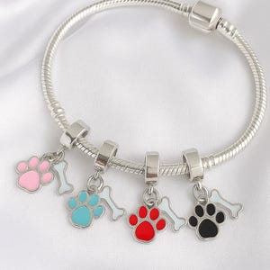 May include: Silver charm bracelet featuring four paw print charms in pink, blue, red, and black, each with a white bone charm. The bracelet has a snake chain design and a barrel clasp.