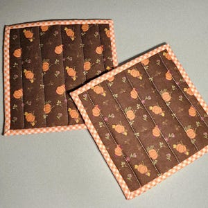 May include: Two square quilted pot holders featuring a brown fabric base with a repeating pattern of orange pumpkins and small green leaves. Each pot holder is finished with an orange and white checkered fabric binding along the edges.