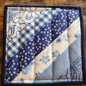 Puede incluir: Posavasos cuadrado acolchado con diseño de patchwork. El posavasos presenta varias telas estampadas en azul y blanco, incluyendo cuadros, flores y estampados de cachemira. Tiene un borde azul oscuro y es para bebidas.