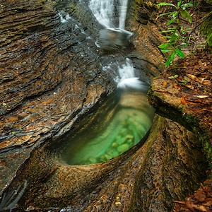 May include: A cascading waterfall flows into a natural pool carved into the rock. The water is clear and green, and the rocks are brown and mossy.