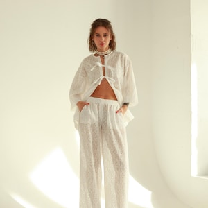 Op de afbeelding: Een wit kanten topje met een strik aan de voorkant en een bijpassende witte kanten broek met wijde pijpen. Het model draagt een zilveren ketting.