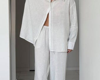 May include: White striped linen pants with a white linen shirt. The pants have a drawstring waist and a lace trim at the bottom. The shirt has a collar and long sleeves.