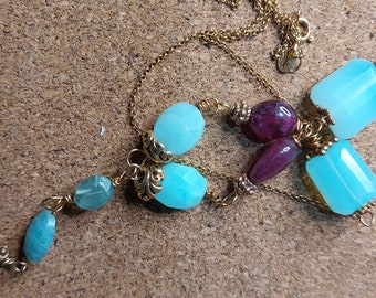Puede incluir: Un collar de cadena dorada con una variedad de cuentas de piedras preciosas azules y moradas. El collar presenta una larga hebra de cuentas colgantes con una piedra preciosa azul más grande en el extremo.