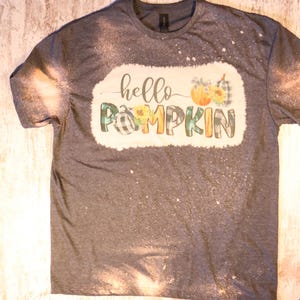 Könnte beinhalten: Ein graues T-Shirt mit einem Bleiche-Spritzer-Design mit dem Text "hello pumpkin" in einem weißen Kasten mit einem grün-schwarzen Karomuster und einer Kürbisgrafik.