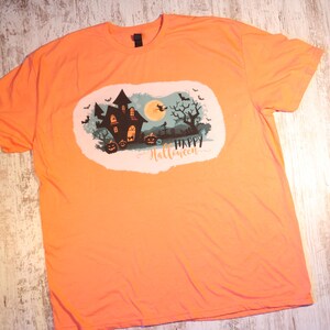 Könnte beinhalten: Orangefarbenes T-Shirt mit einem grafischen Aufdruck eines Spukhauses, eines Vollmonds, Fledermäusen und dem Text "Happy Halloween".