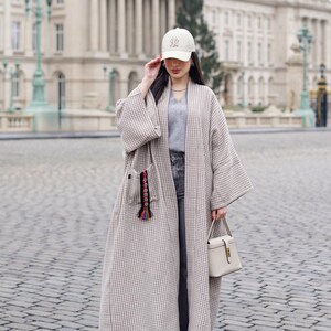 Op de afbeelding: Een lange, open jas in een beige en wit geruit patroon. De jas heeft een zak met kleurrijke kwastjes. De persoon draagt een witte baseballcap met een logo, een grijze top, een jeans en witte sneakers, met een crèmekleurige handtas.