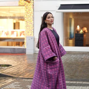 Peut inclure: Long manteau violet à motifs avec bordures et poches blanches. Le manteau présente un motif géométrique et est porté sur un haut noir. L'image est prise à l'extérieur, avec une vitrine et une rue en arrière-plan.