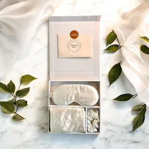 Peut inclure: Coffret cadeau blanc contenant un masque de sommeil, une taie d'oreiller brodée "Aurora" et un chouchou. Une petite enveloppe avec un autocollant circulaire "Merci" est également incluse. Les articles sont disposés sur une surface en marbre.