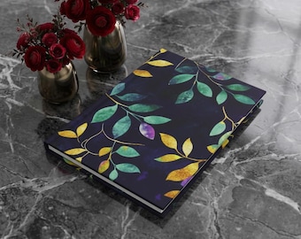 May include: A hardcover journal featuring a dark navy blue cover with a repeating pattern of watercolor-style leaves in shades of green, yellow, and purple. The journal is positioned near two metallic vases filled with deep red ranunculus flowers.