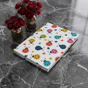 Puede incluir: Un cuaderno blanco con un estampado de mariquitas de colores. Las mariquitas son de varios colores, incluyendo naranja, amarillo, azul y rosa. El cuaderno está sobre una superficie de mármol gris oscuro, con dos jarrones de flores rojas en el fondo.