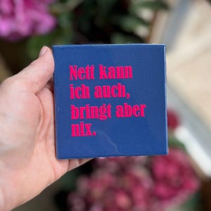Könnte beinhalten: Eine quadratische, glänzend marineblaue Fliese mit dem leuchtend pinken Text "Nett kann ich auch, bringt aber nix." Die Oberfläche ist glatt und reflektierend, wobei die fettgedruckte, serifenlose Schrift deutlich auf der Vorderseite zentriert ist.