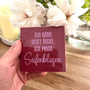 Könnte beinhalten: Eine quadratische, tiefburgunderrote Fliese mit hellrosa Text, der "ICH KANN HEUTE NICHT, ICH MACH Seifenblasen" lautet. Die Fliese hat eine glatte, glänzende Oberfläche und wird in der Hand gehalten, mit weißen Blumen im Hintergrund.