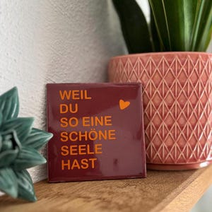 Könnte beinhalten: Eine quadratische, dunkelrote Keramikfliese mit orangefarbenem Text, der "WEIL DU SO EINE SCHÖNE SEELE HAST" lautet, sowie einem kleinen orangefarbenen Herzsymbol. Die Fliese hat eine glatte, glänzende Oberfläche und steht auf einer Holzfläche.