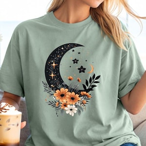 May include: Sage green t-shirt featuring a crescent moon design filled with stars and accented with orange and white flowers. The graphic tee has a relaxed fit and short sleeves, perfect for casual wear.