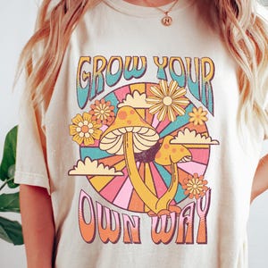 Può includere: Una t-shirt color crema con una grafica retrò di funghi e fiori. Il testo sulla t-shirt recita "Grow Your Own Way".