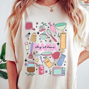 May include: A cream colored t-shirt with a graphic design of kitchen items and the text "stay at home mama".