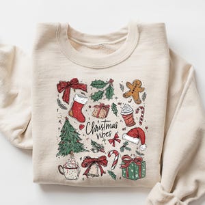 Może przedstawiać: Beżowa bluza z świąteczną grafiką bożonarodzeniową. Projekt zawiera choinkę, skarpetę, piernikowego ludzika i napis "Christmas Vibes". Grafika jest w kolorze czerwonym, zielonym i brązowym.