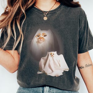 以下が含まれることがあります： 猫の幽霊のデザインが施された濃い灰色のTシャツ。グラフィックは猫の顔と幽霊のコスチュームを着た猫を示しています。このTシャツは快適でカジュアルなスタイルです。