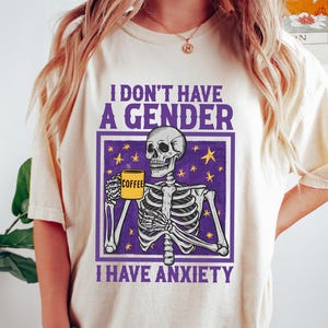 Peut inclure: T-shirt crème avec un motif de squelette tenant une tasse à café. Le texte dit "I DON'T HAVE A GENDER, I HAVE ANXIETY" en violet et blanc. Le squelette est dans un cadre violet avec des étoiles jaunes.