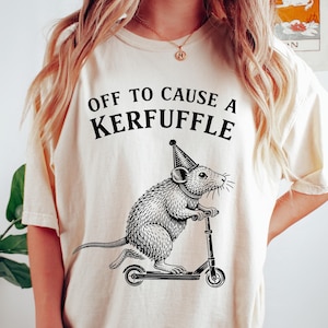 Peut inclure: T-shirt crème avec l'inscription "OFF TO CAUSE A KERFUFFLE" surmontant une illustration en noir et blanc d'un rat sur une trottinette, portant un chapeau de fête. Le t-shirt est de coupe ample.