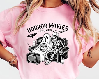 May include: Pink t-shirt with the text "HORROR MOVIES AND CHILL" above a graphic of a ghost and skeleton watching TV. The ghost and skeleton are seated on a couch with popcorn, a bottle, and bats.