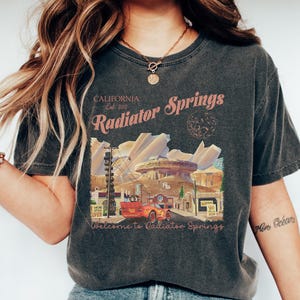 以下が含まれることがあります： 「CALIFORNIA Est. 202」と「Radiator Springs」の文字と、街並みを背景にヴィンテージカーが描かれたダークグレーのTシャツ。「Welcome to Radiator Springs」の文字も含まれています。