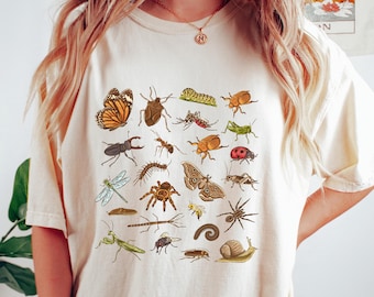 May include: Cream-colored t-shirt featuring a colorful insect illustration. The design includes various bugs like butterflies, beetles, and spiders. The shirt has a relaxed fit and short sleeves, perfect for casual wear.
