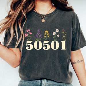Peut inclure: T-shirt gris foncé avec des motifs floraux au-dessus des chiffres "50501" en crème. Le t-shirt est porté avec un jean bleu et des baskets blanches. L'ensemble est décontracté et confortable.