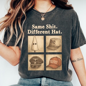 Könnte beinhalten: Dunkelgraues T-Shirt mit dem Text "Same Shit. Different Hat." und Illustrationen verschiedener Hüte. Die Hüte umfassen eine KKK-Haube, einen Militärhut und einen "Make America Great Again"-Hut.