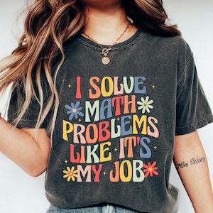 May include: Dark grey t-shirt with the slogan "I SOLVE MATH PROBLEMS LIKE IT'S MY JOB" in colourful, retro lettering. The shirt is worn with blue jeans and white trainers. The theme is math and education.