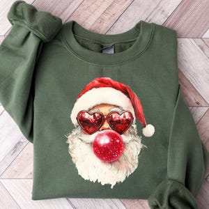Op de afbeelding: Een olijfgroene sweatshirt met een afbeelding van de Kerstman met hartvormige zonnebrillen en een rode kauwgomballenbel. De Kerstman draagt een rood-witte muts. De sweatshirt heeft lange mouwen met geribde manchetten.