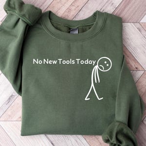 Könnte beinhalten: Ein olivgrünes Sweatshirt mit dem Text "No New Tools Today" und einer traurigen Strichmännchen-Grafik in Weiß. Das Sweatshirt hat einen Rundhalsausschnitt und lange Ärmel mit gerippten Bündchen. Der Hintergrund ist eine helle Holzoberfläche.